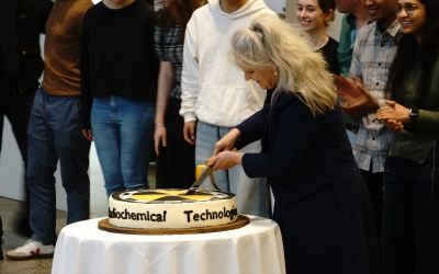 A group of people gathered around a table with a cake that has "Biochemical Technology" written on its side. One person with long hair is cutting the cake, while others are standing nearby, watching and celebrating.