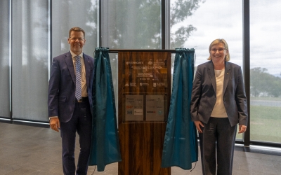 Mr Tony Cook, Secretary of the Department of Education and Professor Ute Roessner, CEO of the Australian Research Council stand either side of a wooden plaque on a stand with teal curtains. The plaque displays engraved text and logos. The setting is indoors with large windows with and a view of trees outside.