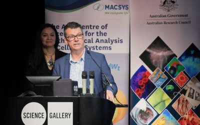 Director of the ARC Centre of Excellence for the Mathematical Analysis of Cellular Systems (MACSYS) Professor Trevor Lithgow stands at a lectern labelled "SCIENCE GALLERY" delivering a speech or presentation. Trevor is wearing a patterned shirt and a blue jacket, positioned behind two microphones and a computer monitor. In the background, MC Tanya Ha is partially visible, standing slightly behind Trevor. Behind both individuals are banners for MACSYS.