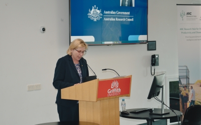 A speaker stands at a podium bearing the Griffith University logo, delivering a presentation. Behind them, a large screen displays the Australian Government and Australian Research Council logos, along with an image of another individual. To the right, a banner for the ARC Research Hub for Driving Farming Productivity and Disease Prevention is visible. The setting appears to be a formal conference or academic event, with a computer monitor and water bottle on a nearby table.