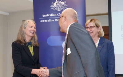 Two individuals shaking hands in front of a banner that reads: ‘Australian Government Australian Research Council.’ A third person stands nearby. All are wearing formal attire and name tags.