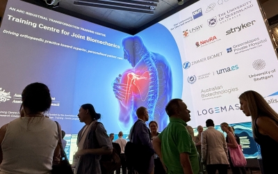 Attendees networking inside QUT’s The Cube, with a large digital display showcasing the ARC Industrial Training Centre for Joint Biomechanics and its partner organisations.