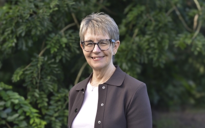 Prof Janeen Baxter wearing glasses, a light‑coloured top and a brown jacket, standing outdoors with green foliage behind them