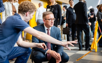 The Hon. Julian Hill MP, Assistant Minister for Citizenship, Customs and Multicultural Affairs and Assistant Minister for International Education, crouches beside a robotics demonstration while another person gestures towards the equipment, with other event participants in the background.