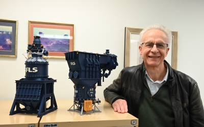 Laureate Professor Kevin Galvin standing beside two scale models of industrial mining equipment displayed on a cabinet in an office setting, with framed landscape photographs in the background.