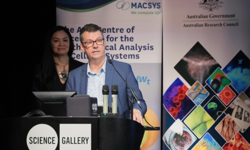 Director of the ARC Centre of Excellence for the Mathematical Analysis of Cellular Systems (MACSYS) Professor Trevor Lithgow stands at a lectern labelled "SCIENCE GALLERY" delivering a speech or presentation. Trevor is wearing a patterned shirt and a blue jacket, positioned behind two microphones and a computer monitor. In the background, MC Tanya Ha is partially visible, standing slightly behind Trevor. Behind both individuals are banners for MACSYS.