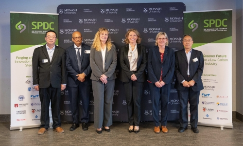 Monash VC and President Prof Sharon Pickering, ARC CEO Prof Ute Roessner and Hub leadership standing in a row in front of a backdrop with Monash University branding. Two banners on either side display the Hub logo and text about forging a greener future for a low-carbon steel industry.