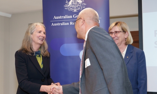 Two individuals shaking hands in front of a banner that reads: ‘Australian Government Australian Research Council.’ A third person stands nearby. All are wearing formal attire and name tags.