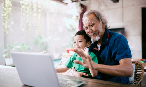 An older person and a child sitting together at a wooden table, looking at a laptop screen. The older person is wearing a dark blue shirt with red trim, and the child is in a green shirt. They appear to be engaged in a video call, with the older person raising a hand in a gesture toward the laptop. The setting is a bright indoor space with natural light, plants in the background, and a concrete wall.