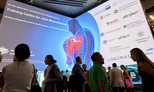 Attendees networking inside QUT’s The Cube, with a large digital display showcasing the ARC Industrial Training Centre for Joint Biomechanics and its partner organisations.