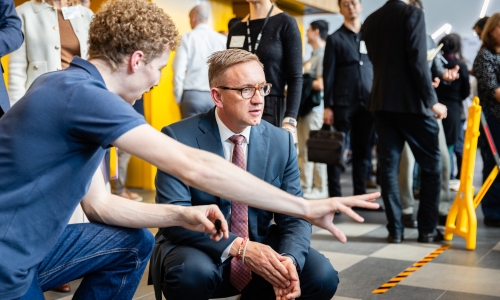 The Hon. Julian Hill MP, Assistant Minister for Citizenship, Customs and Multicultural Affairs and Assistant Minister for International Education, crouches beside a robotics demonstration while another person gestures towards the equipment, with other event participants in the background.