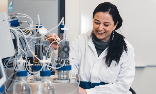 Person in a white laboratory coat operating analytical laboratory equipment connected with multiple tubes and containers. Several clear bottles are positioned in the foreground, and the laboratory instrument occupies much of the left side of the image.