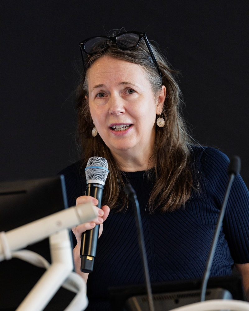 ARC Academic Director Prof Alison Ross holding a microphone while standing near a podium with visible equipment. She is wearing a dark top and has glasses resting on her head. The background is dark.