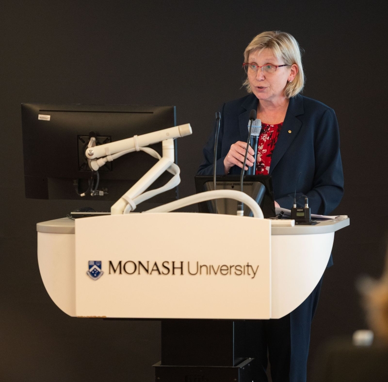 ARC CEO Prof Ute Roessner standing at a podium with the Monash University logo, holding a microphone. A computer monitor and presentation equipment are visible on the podium.