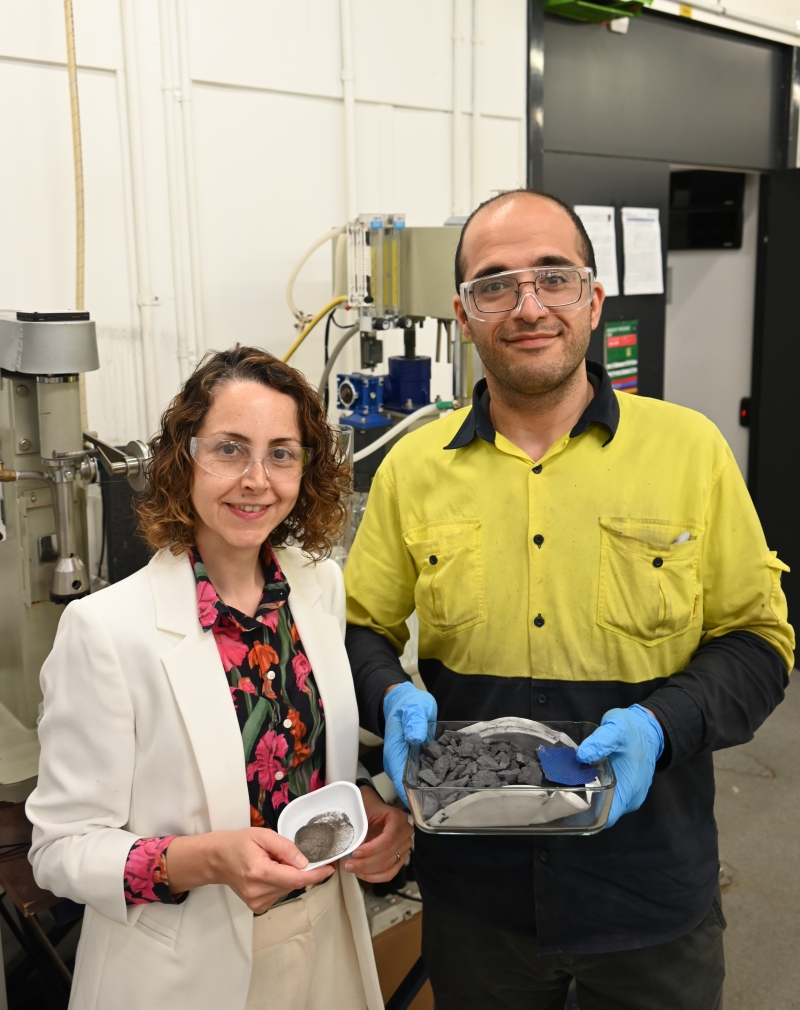 Associate Professor Mahshid Firouzi and PhD candidate Hamidreza Saffarian people wearing safety glasses stand in an industrial laboratory beside processing equipment, holding trays containing powdered and rock-like mineral samples.