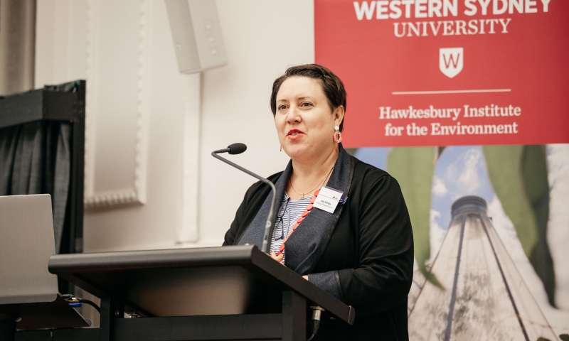 Ms Judy Satrapa stands at a podium delivering a speech, with a banner in the background displaying