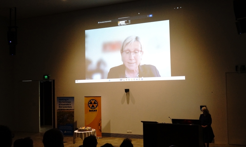 A large screen in a dimly lit room shows a virtual meeting with ARC CEO Ute Roessner. The image of Ute Roessner is projected on the screen, and her face is obscured by a pink square. Below the screen, there are two banners; and a person stands at a podium to the right side of the room.