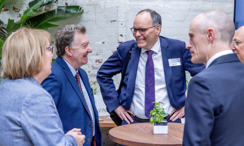 Professor Chris Pakes, Deputy Vice-Chancellor (Research & Industry Engagement) at La Trobe University (right), and Professor Alastair McEwan, ARC Executive Director, Biological Sciences and Biotechnology (left), standing and conversing with others around a small round table with a potted plant, at the ARC Research Hub for Protected Cropping (PC Hub) launch event. The setting includes a brick wall and leafy indoor plants in the background.
