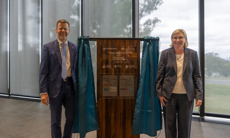Mr Tony Cook, Secretary of the Department of Education and Professor Ute Roessner, CEO of the Australian Research Council stand either side of a wooden plaque on a stand with teal curtains. The plaque displays engraved text and logos. The setting is indoors with large windows with and a view of trees outside.
