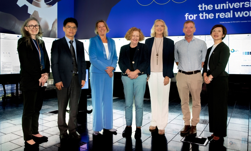 Prof Jennifer Macleod, Hub Director Prof Zhigang Chen, ARC Academic Director Prof Anika Gauja, QUT Vice Chancellor Prof Margaret Sheil, Ms Madonna Jarrett MP, CEO of Halocell Energy Paul Moonie, and Hub Deputy Director Prof Hongxia Wang standing in a row inside the event venue. Behind them is a large blue display with text “the university for the real world” and diagrams, as well as scientific posters.