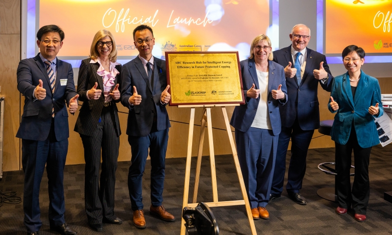 A group of seven individuals including ARC CEO Prof Ute Roessner standing together indoors, all giving thumbs-up gestures. They are positioned around a wooden easel holding a plaque. A large screen in the background displays the words 