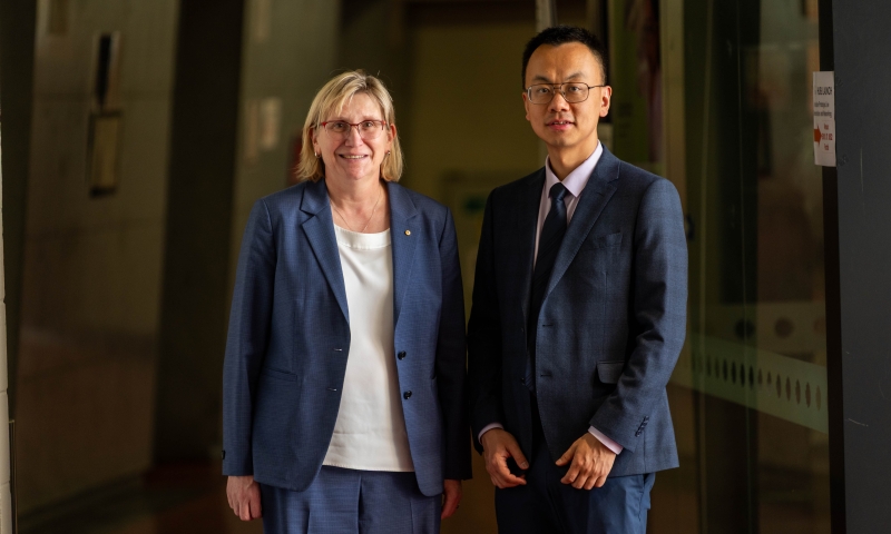 ARC CEO Prof Ute Roessner and Hub Director Distinguished Prof Tianyi Ma standing side by side in a hallway, both wearing formal business suits. The background shows a corridor with muted lighting and glass panels on one side.