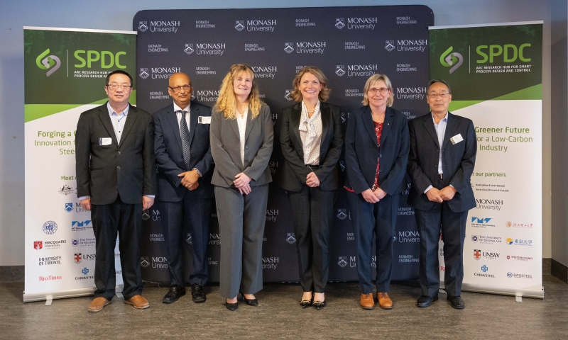 Monash VC and President Prof Sharon Pickering, ARC CEO Prof Ute Roessner and Hub leadership standing in a row in front of a backdrop with Monash University branding. Two banners on either side display the Hub logo and text about forging a greener future for a low-carbon steel industry.