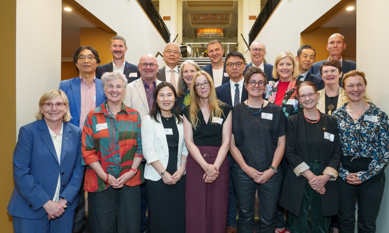 A group photo of people standing on a staircase in a formal setting. They are wearing business attire and name tags. The background includes railings and a decorative hallway.