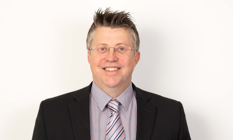 Person, identified as Mr Craig Maconachie, wearing a black suit jacket over a light-colored shirt with a striped tie, posed against a plain white background.