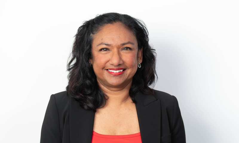 Person, identified as Ms Geraldine Dennis, wearing a black blazer over a bright red top, with wavy shoulder-length hair, posed against a plain white background.