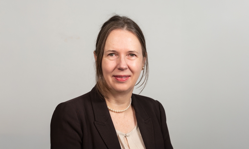 Person, identified as Prof Alison Ross, wearing a dark blazer over a light blouse, accessorised with a pearl necklace, posed against a plain light gray background.