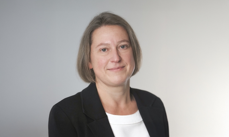 Person, identified as Prof Anika Gauja, wearing a black blazer over a white top, posed against a light gray background.