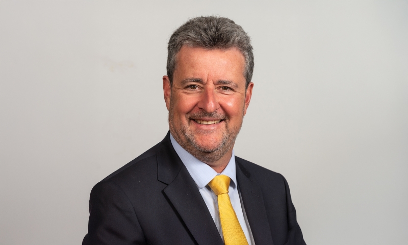 Person, identified as Prof Steven Weller, wearing a dark suit jacket, light blue dress shirt, and a bright yellow tie, posed against a plain light gray background.