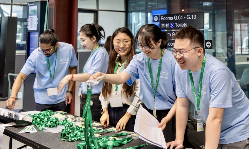 Five people standing behind a table, wearing light blue shirts and green lanyards, and handing out green lanyards to attendees.