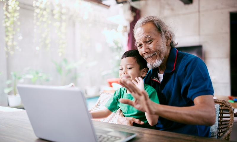 An older person and a child sitting together at a wooden table, looking at a laptop screen. The older person is wearing a dark blue shirt with red trim, and the child is in a green shirt. They appear to be engaged in a video call, with the older person raising a hand in a gesture toward the laptop. The setting is a bright indoor space with natural light, plants in the background, and a concrete wall.