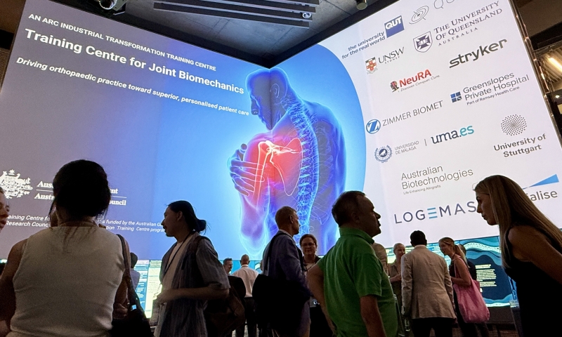 Attendees networking inside QUT’s The Cube, with a large digital display showcasing the ARC Industrial Training Centre for Joint Biomechanics and its partner organisations.