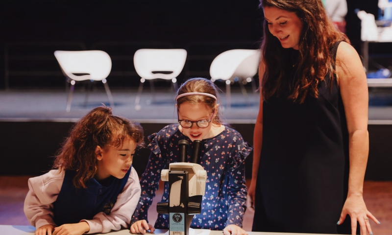 Two primary school-aged students examine a microscope on a table while an adult facilitator stands beside them at a science outreach event, with learning materials spread out in front.
