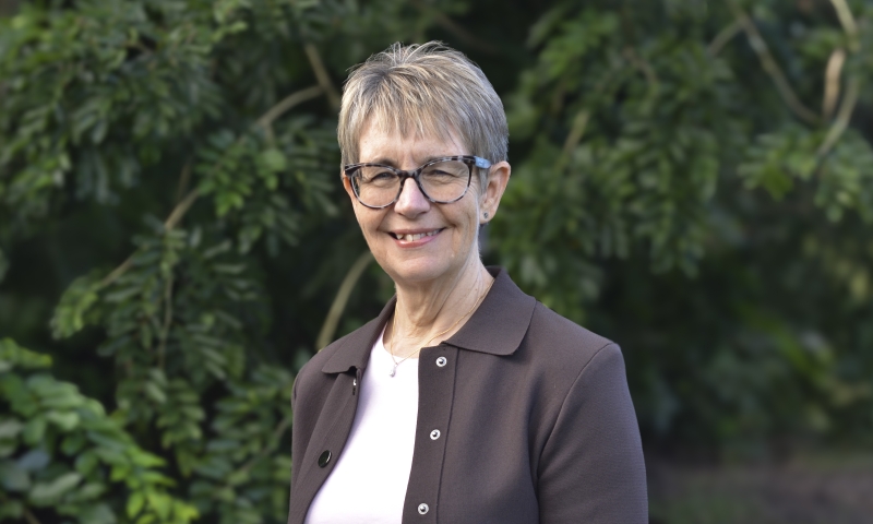 Prof Janeen Baxter wearing glasses, a light‑coloured top and a brown jacket, standing outdoors with green foliage behind them