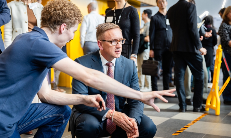 The Hon. Julian Hill MP, Assistant Minister for Citizenship, Customs and Multicultural Affairs and Assistant Minister for International Education, crouches beside a robotics demonstration while another person gestures towards the equipment, with other event participants in the background.