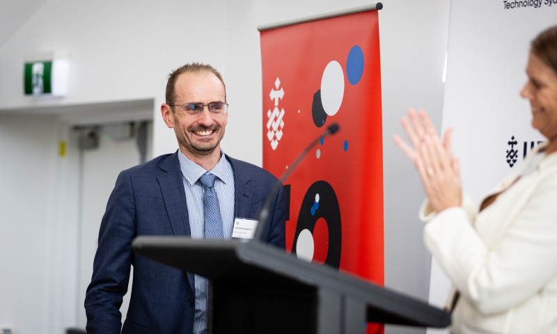 ARC Deputy Chief Executive Officer Anthony Murfett stands left of stage waiting to join the stage during the HaRTCon launch at the University of Technology Sydney, with event branding visible behind him and an attendee applauding nearby.