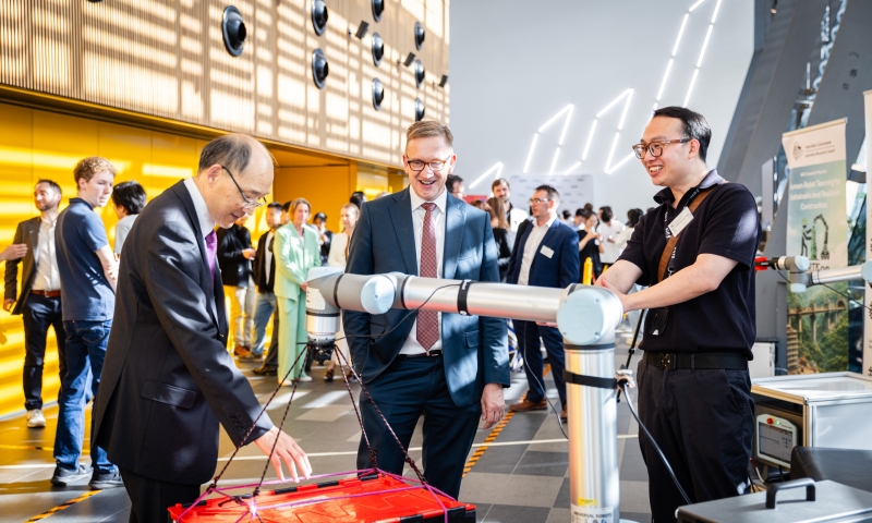 A collaborative robotic arm lifts a toolbox during a demonstration at the HaRTCon launch, observed by the Hon. Julian Hill MP and Distinguished Professor Dikai Liu, with other attendees in the background.