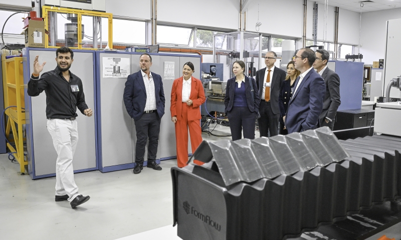 A presenter gestures while speaking to a small group during a tour inside a manufacturing or research facility, with equipment, control panels and industrial machines surrounding them.