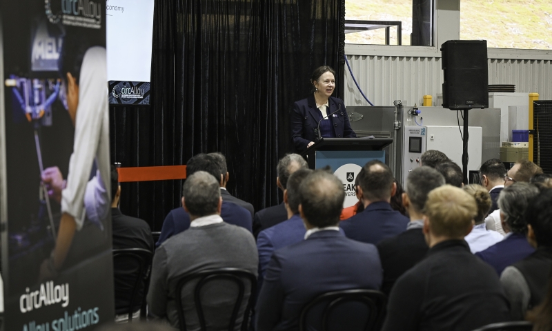 Prof Alison Ross stands at a lectern addressing a seated audience in an indoor event space, with banners and a screen displaying 