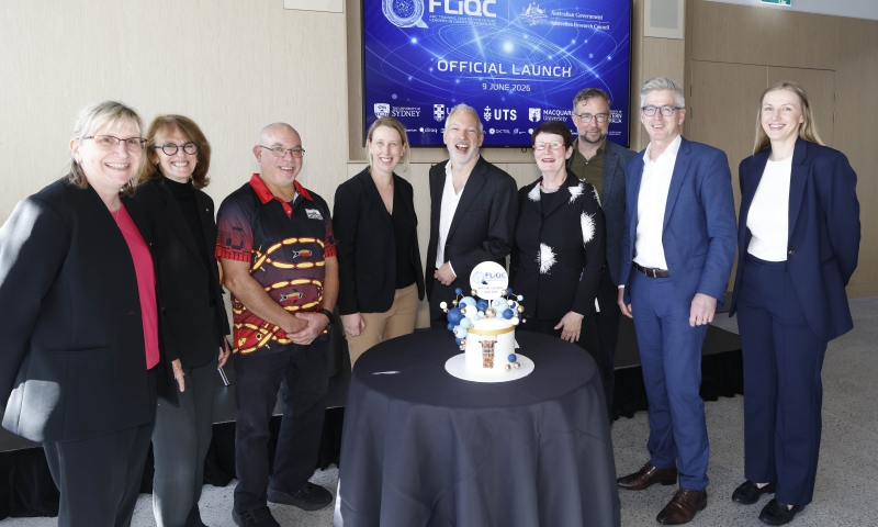 A group of people standing around a table with a cake in front of a screen displaying 