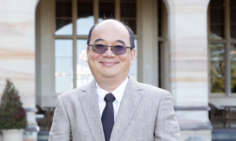 Portrait of a researcher wearing glasses, a light grey suit and dark tie, standing in front of a building with arched windows.